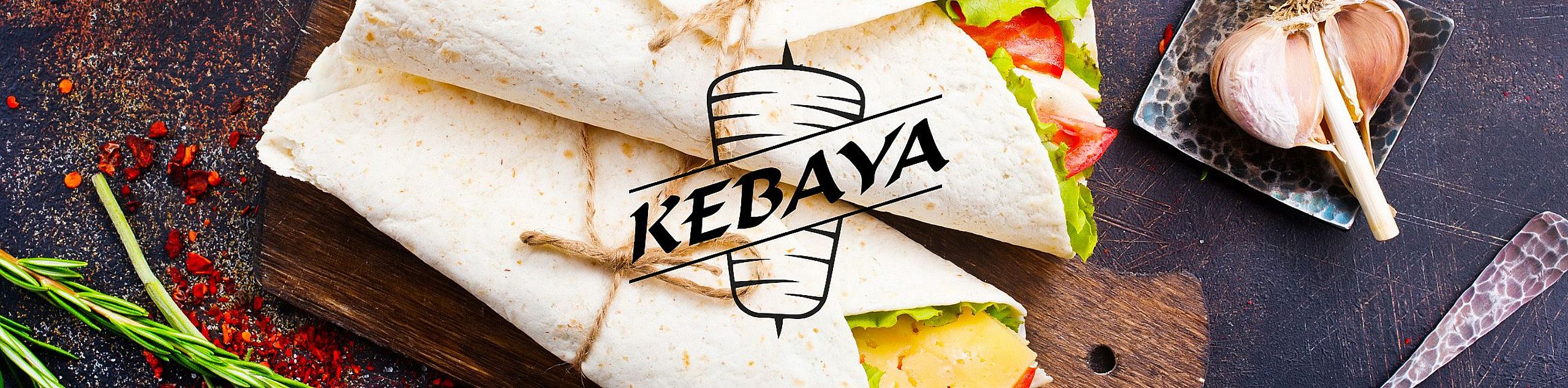 Zbliżenie na apetyczne wrapy w tortilli wypełnione świeżymi warzywami i serem, starannie związane sznurkiem i ułożone na drewnianej desce. W tle przyprawy, czosnek i gałązka rozmarynu. Na środku logo „KEBAYA” z graficznym motywem kebaba.