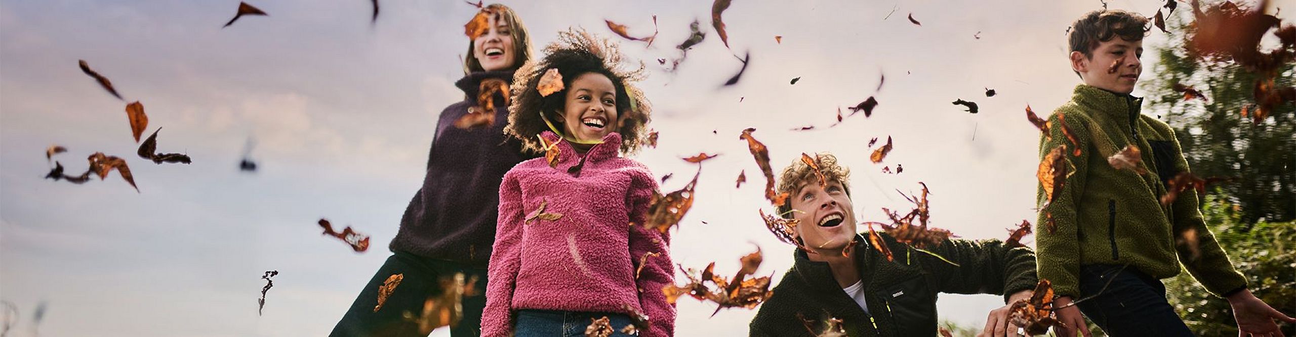 Grupa dzieci i dorosłych bawi się wśród opadających jesiennych liści na świeżym powietrzu. Wszyscy uśmiechnięci, ubrani w ciepłe, kolorowe polary – w odcieniach różu, zieleni i ciemnego fioletu. Tło stanowi pastelowe niebo i jesienna roślinność.