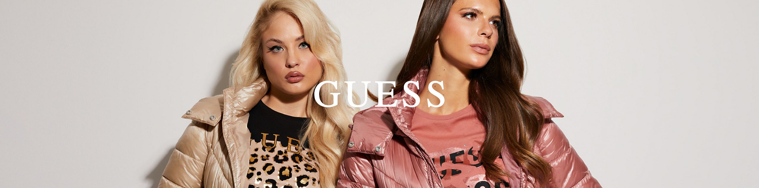 Dwie kobiety pozują na tle białej ściany w błyszczących, pikowanych kurtkach GUESS — jedna w jasnobeżowej z cętkowanym topem, druga w pudrowo-różowej z różową koszulką z nadrukiem. Na środku znajduje się białe logo „GUESS”.