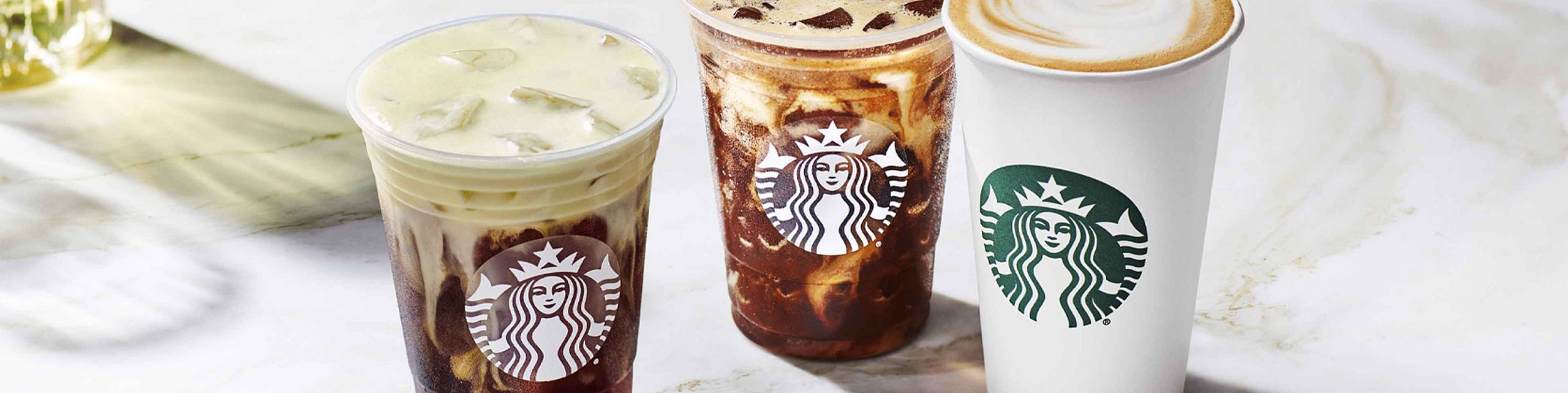 Three Starbucks beverages placed on a marble surface – two iced drinks in plastic cups (one light, one dark) and one hot drink in a white paper cup with milk foam.