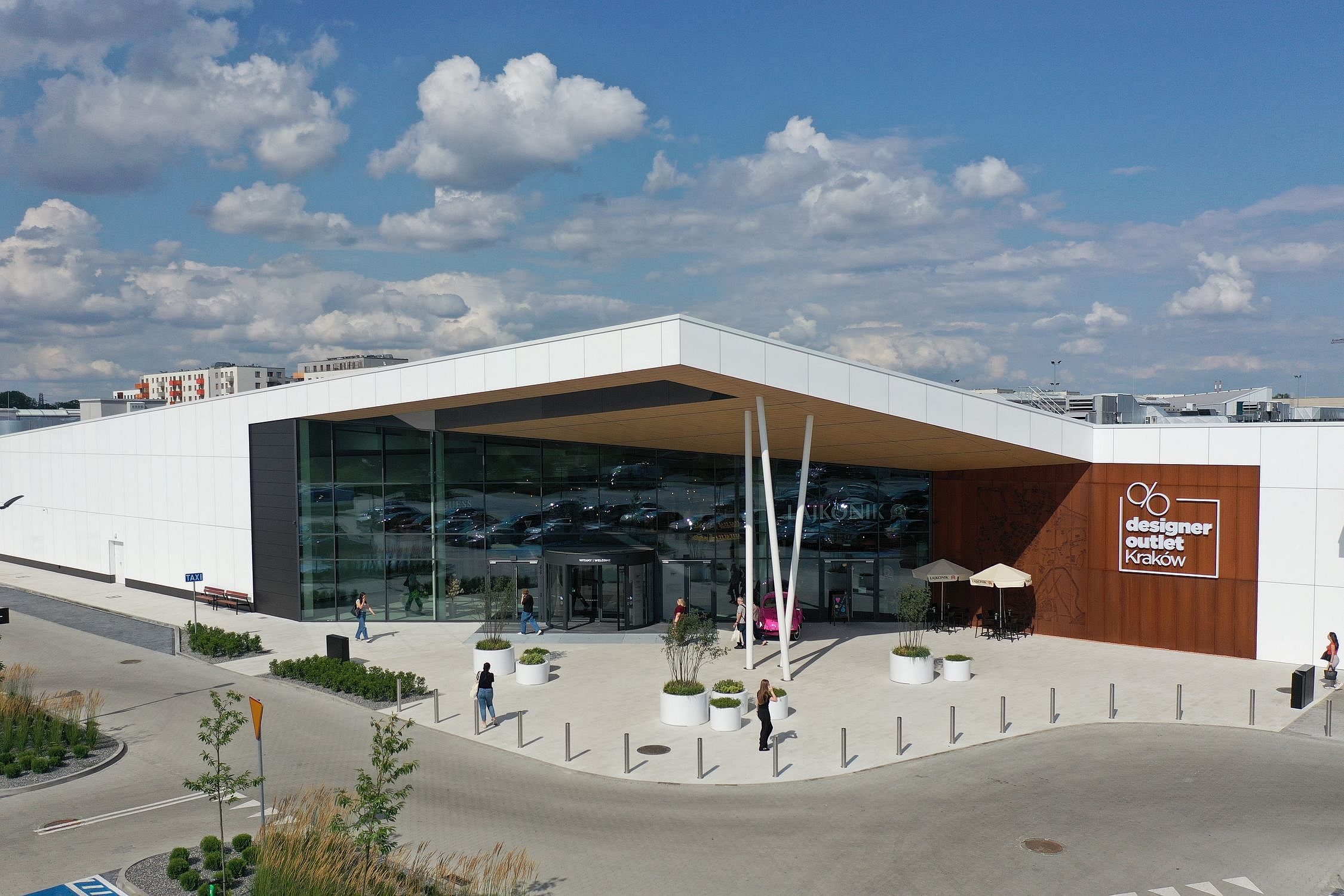 Frontowe wejście do Designer Outlet Kraków z nowoczesną architekturą i dużymi przeszklonymi ścianami. Nad drzwiami obrotowymi widoczny jest napis "Witamy / Welcome". Po lewej stronie znak TAXI, po prawej logo outletu oraz kawiarnia La Konik z różowym samochodem retro i parasolami. Przed wejściem ludzie siedzą i spacerują.