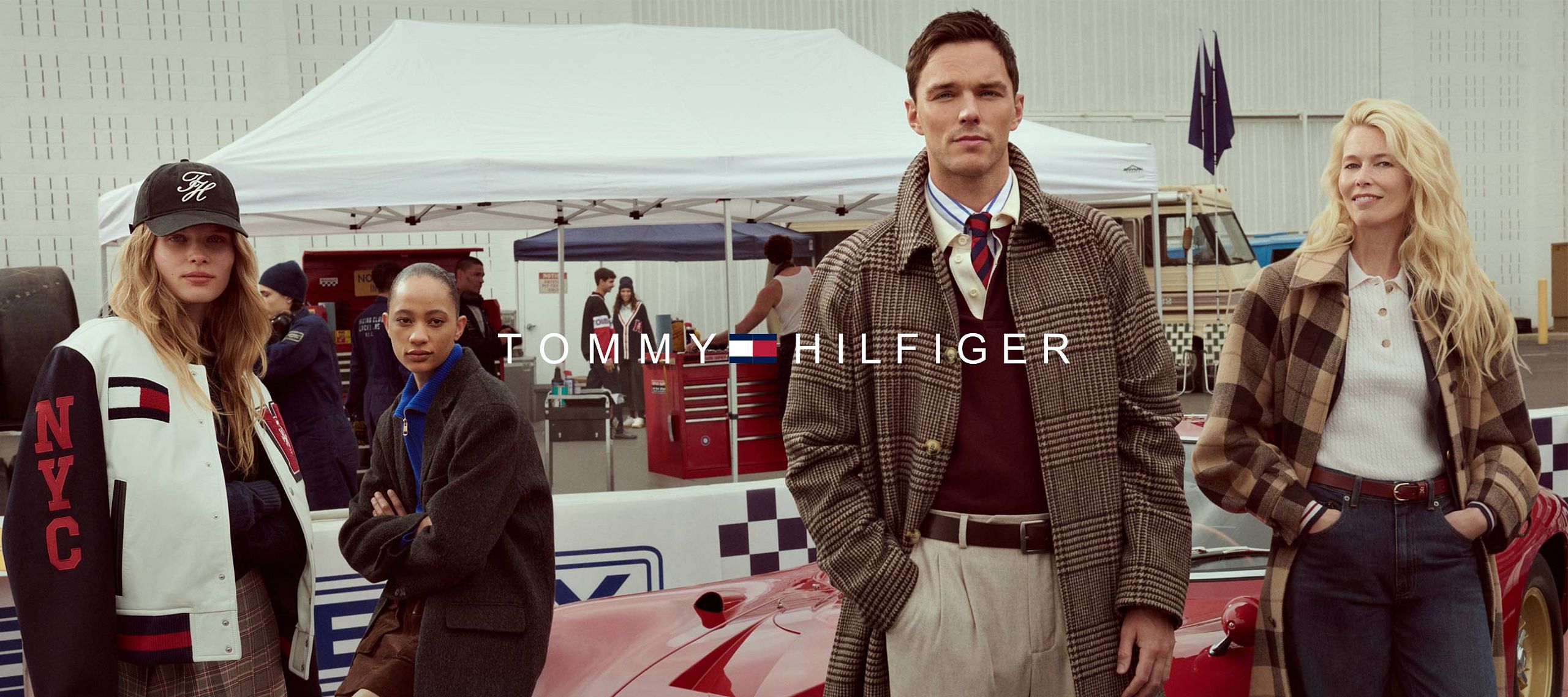 Grupa osób w stylowych ubraniach w klimacie retro stoi przy czerwonym samochodzie wyścigowym. W tle widoczny jest namiot serwisowy i logo Tommy Hilfiger na środku obrazu.