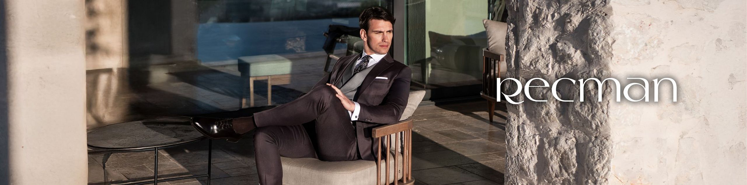 A man in an elegant Recman suit sits on a terrace beside a stone wall, with a modern glass-door interior in the background.