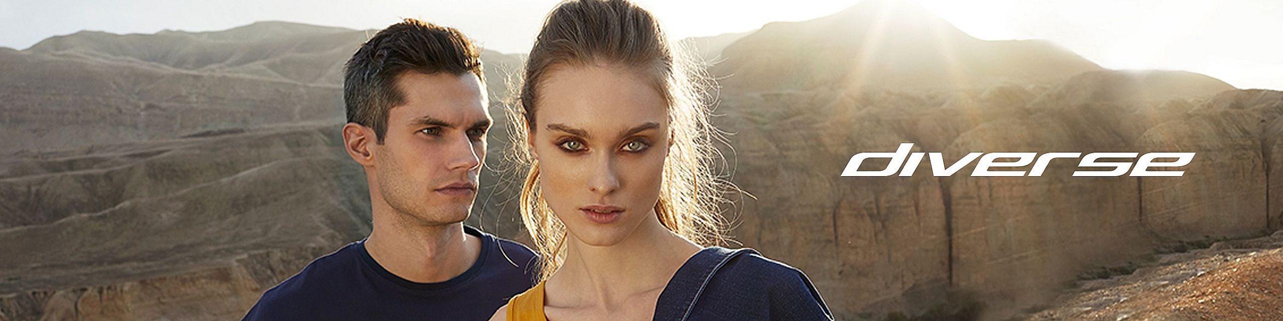 A man and a woman confidently look ahead, posing in Diverse clothing against a rocky desert landscape illuminated by the setting sun. On the right side appears the white “diverse” logo.