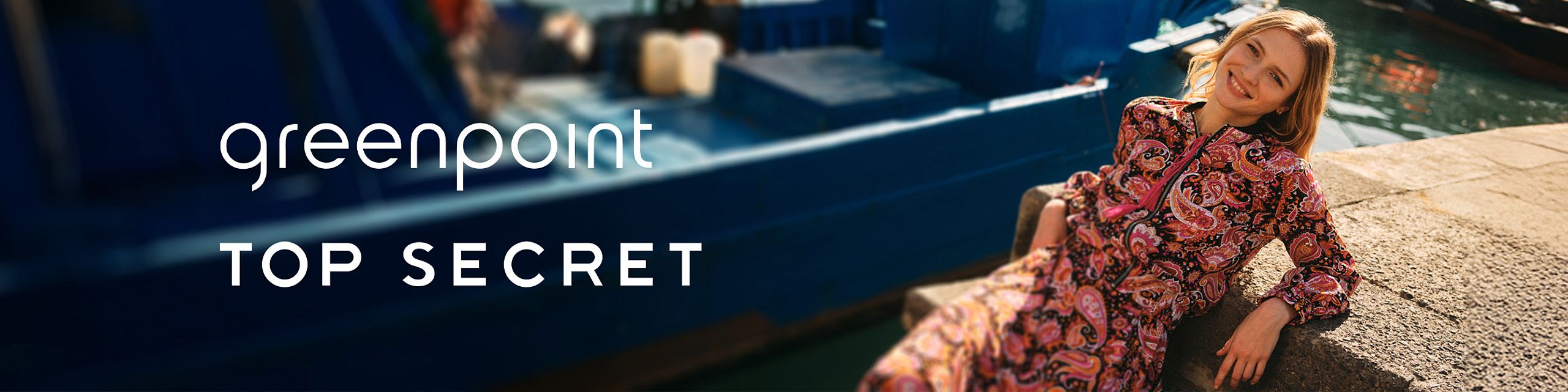 Kobieta w kolorowej sukience z motywem paisley uśmiecha się, siedząc na nabrzeżu przy wodzie. W tle widoczna niebieska łódź. Logo marek Greenpoint i Top Secret.