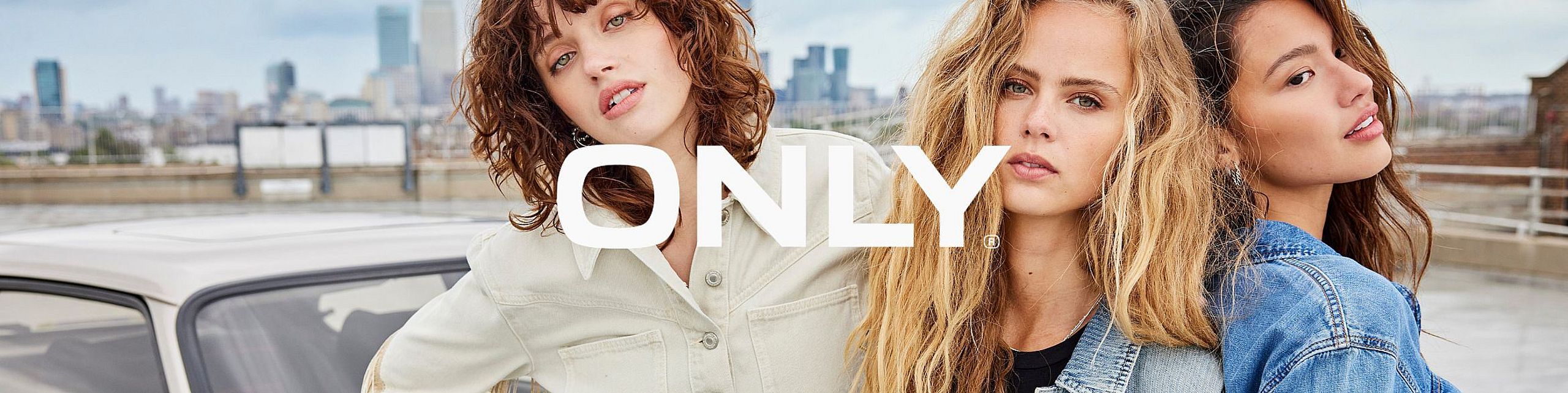 Trzy młode kobiety pozują razem na tle miejskiej panoramy, ubrane w casualowe stylizacje marki ONLY – m.in. jeansowe i kremowe kurtki. Na środku widoczne duże, białe logo „ONLY”.