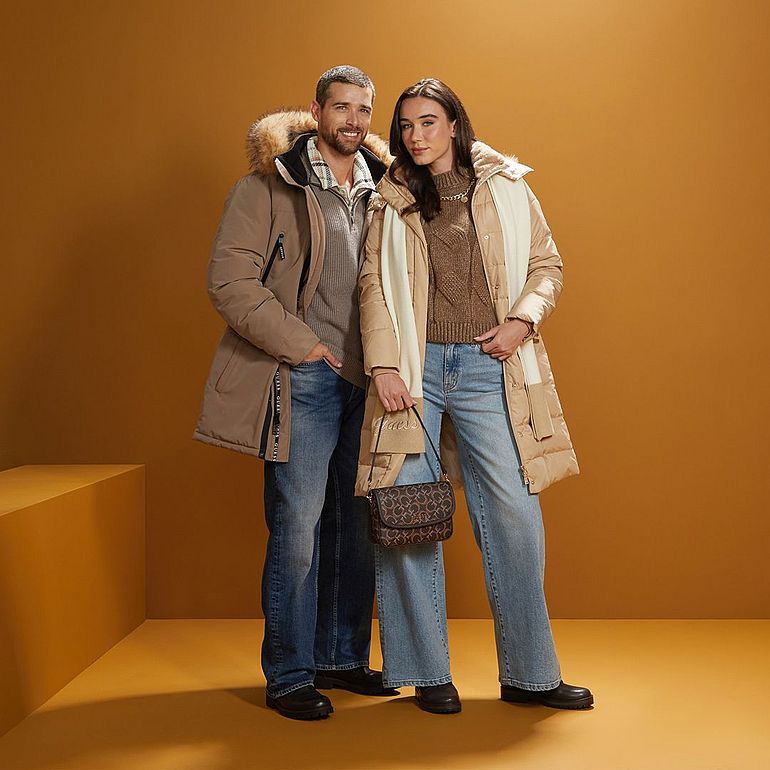 A couple dressed in winter outfits, wearing beige down jackets and jeans, poses against a plain orange background.