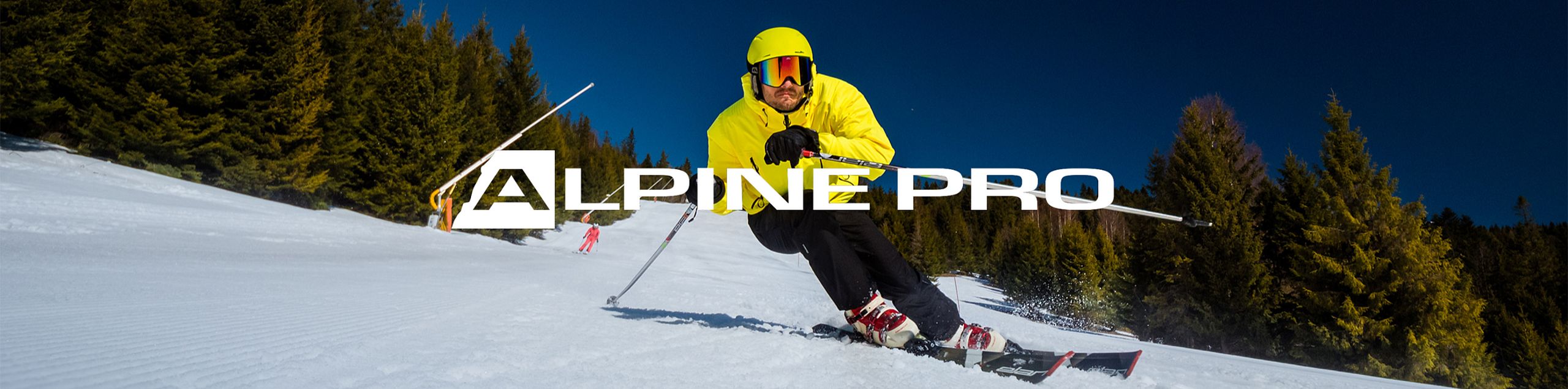 A skier in a yellow jacket descends a snowy slope surrounded by forest, under a clear blue sky. The "ALPINE PRO" logo is prominently displayed in the center.