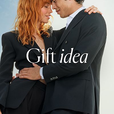 A couple dressed in elegant suits, standing close together and gazing into each other's eyes in a romantic embrace. In the background, the bright sky shows the text "Gift Idea".
