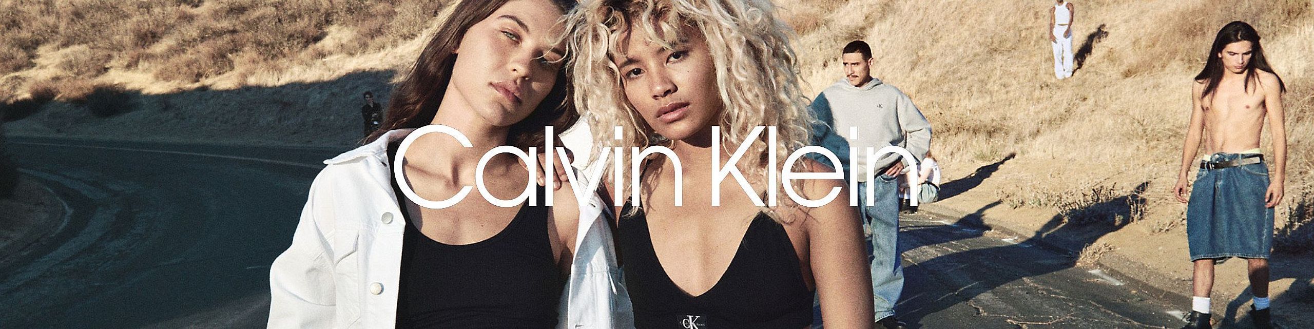 Na tle pustynnego krajobrazu dwie młode osoby stoją blisko siebie – jedna z głową opartą na ramieniu drugiej. Oboje ubrani są w proste, casualowe ubrania marki Calvin Klein. W tle widać inne osoby w podobnym stylu. Na środku umieszczono biały napis „Calvin Klein”.