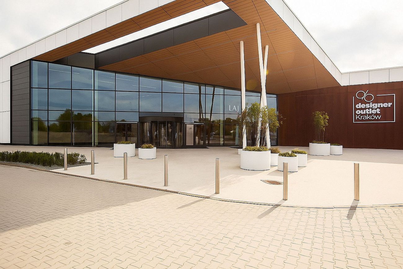 Nowoczesne wejście do centrum handlowego Designer Outlet Kraków, z przeszkloną fasadą, donicami z roślinami i widocznym logo obiektu po prawej stronie.