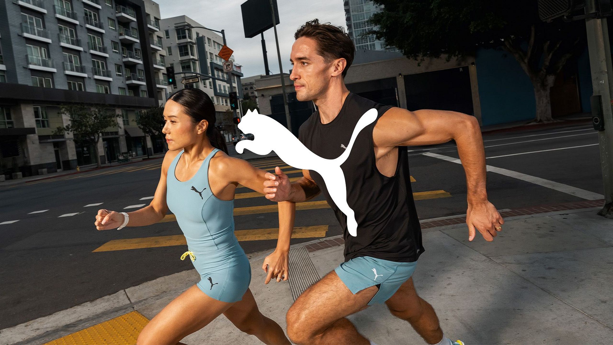 Kobieta i mężczyzna biegną ulicą w odzieży sportowej PUMA. Na zdjęciu widoczne duże logo marki.