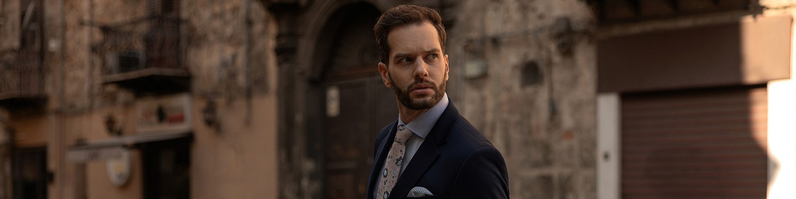 Elegancki mężczyzna w granatowym garniturze i krawacie w delikatny wzór, spacerujący po klimatycznej, kamiennej uliczce miasta – stylowa moda męska VILLARO.