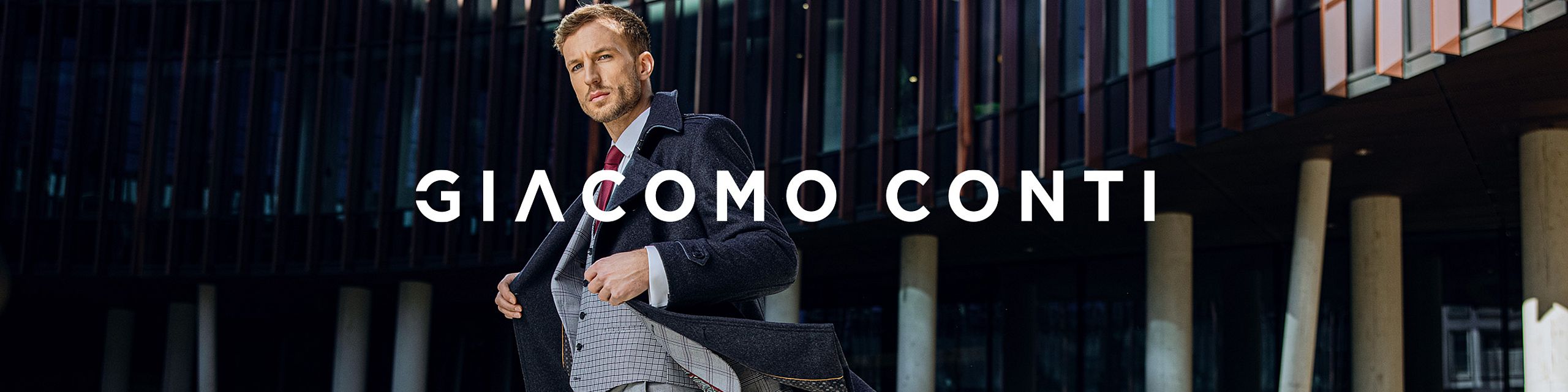 Elegancki mężczyzna w granatowym płaszczu i klasycznym garniturze z czerwonym krawatem poprawia swoją marynarkę, stojąc na tle nowoczesnego budynku z przeszkloną fasadą. Na środku widoczne jest białe logo „GIACOMO CONTI”.
