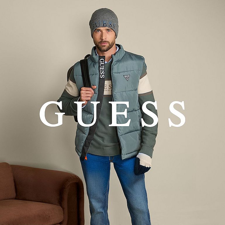 Mężczyzna w zimowej stylizacji marki Guess: szara czapka z logo, niebieska pikowana kamizelka, sweter w odcieniach zieleni i beżu, jeansy oraz torba na ramię; stoi na tle beżowej ściany obok brązowej kanapy. Na środku widoczne duże logo GUESS.