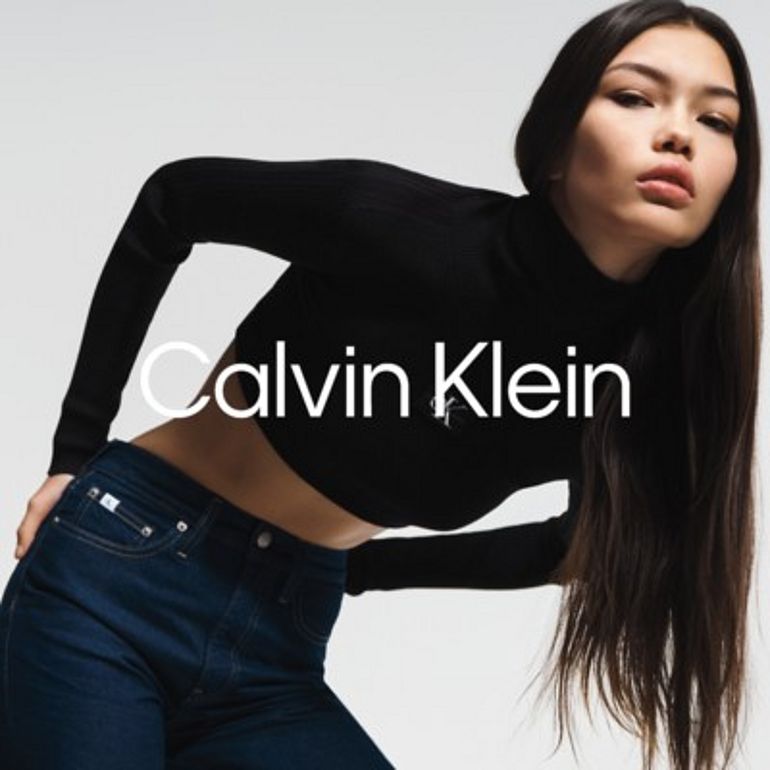 Modelka w czarnym golfie i jeansach marki Calvin Klein pozuje na jasnym tle. Na zdjęciu widoczne logo Calvin Klein.