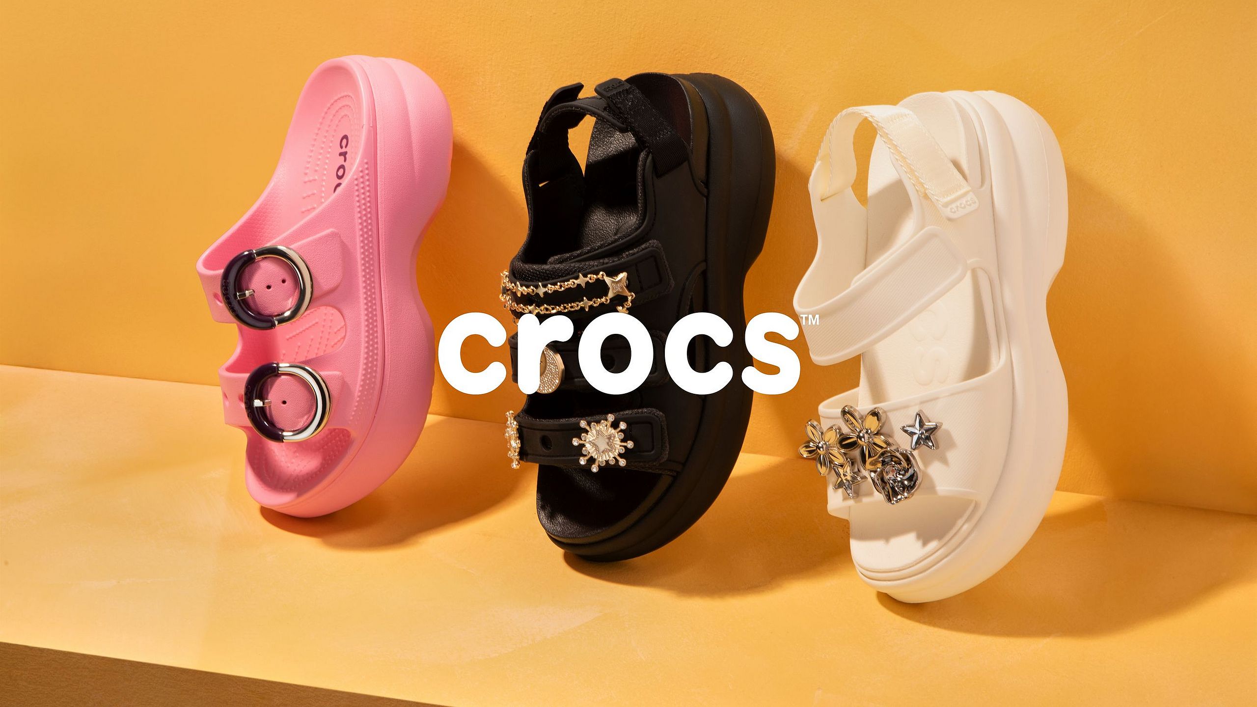 Trzy modele sandałów Crocs w kolorach różowym, czarnym i kremowym na pomarańczowym tle z logo marki.