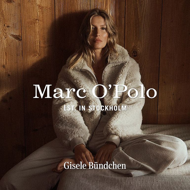 Modelka siedząca w jasnym, futrzanym stroju na tle drewnianej ściany; reklama marki Marc O’Polo.