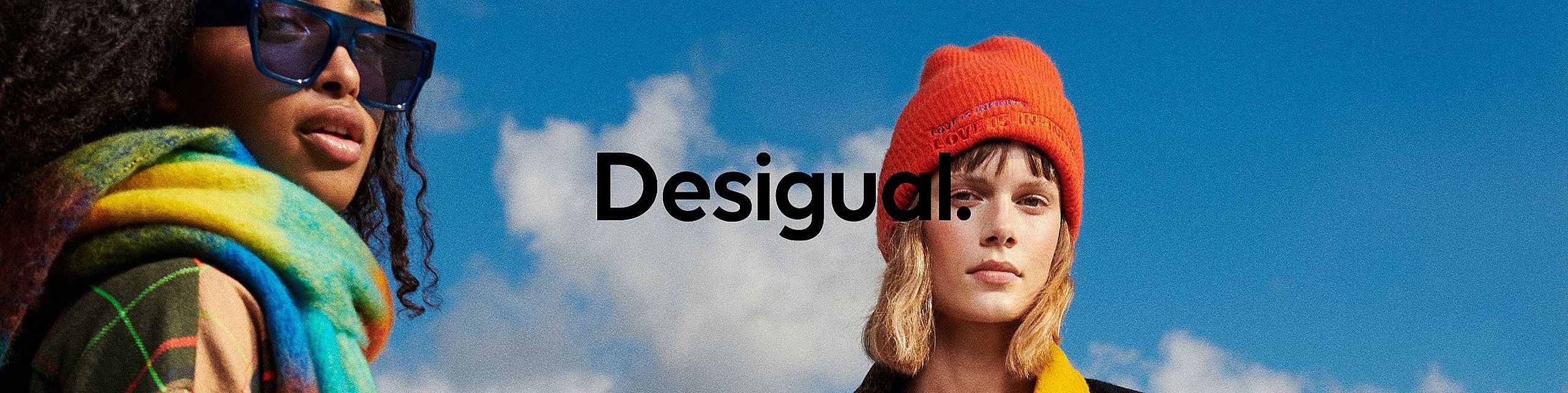 Dwie młode osoby na tle nieba z białymi chmurami. Po lewej modelka z kręconymi włosami, w dużych granatowych okularach przeciwsłonecznych i kolorowym, tęczowym szalu. Po prawej osoba w czerwonej czapce beanie z napisem i żółtej kurtce. Pośrodku, na tle nieba, czarne logo marki Desigual.