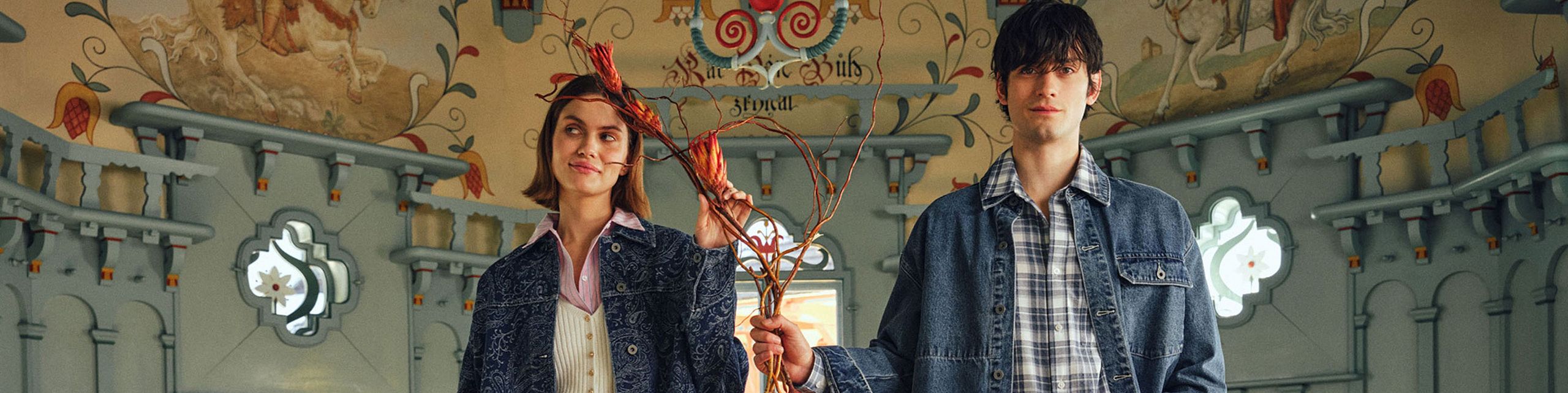 Two young people holding branches inside an ornate painted hall.