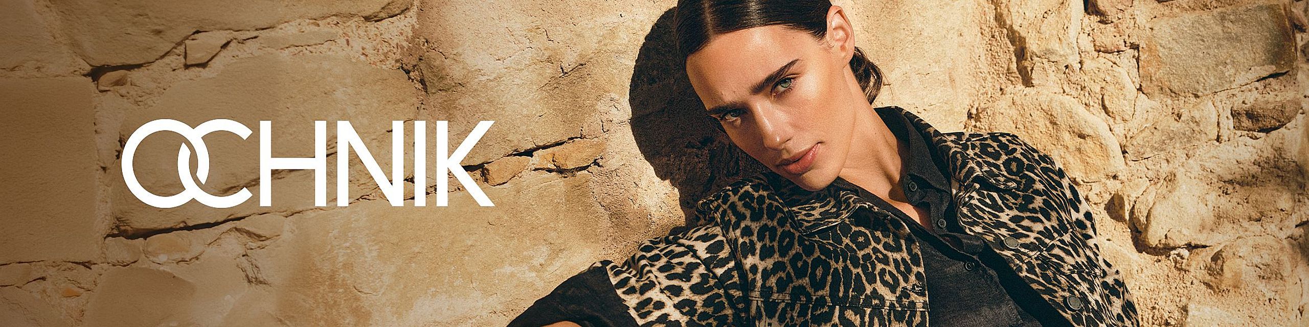 Modelka w kurtce w panterkę pozuje na tle kamiennej ściany, obok logo marki OCHNIK.