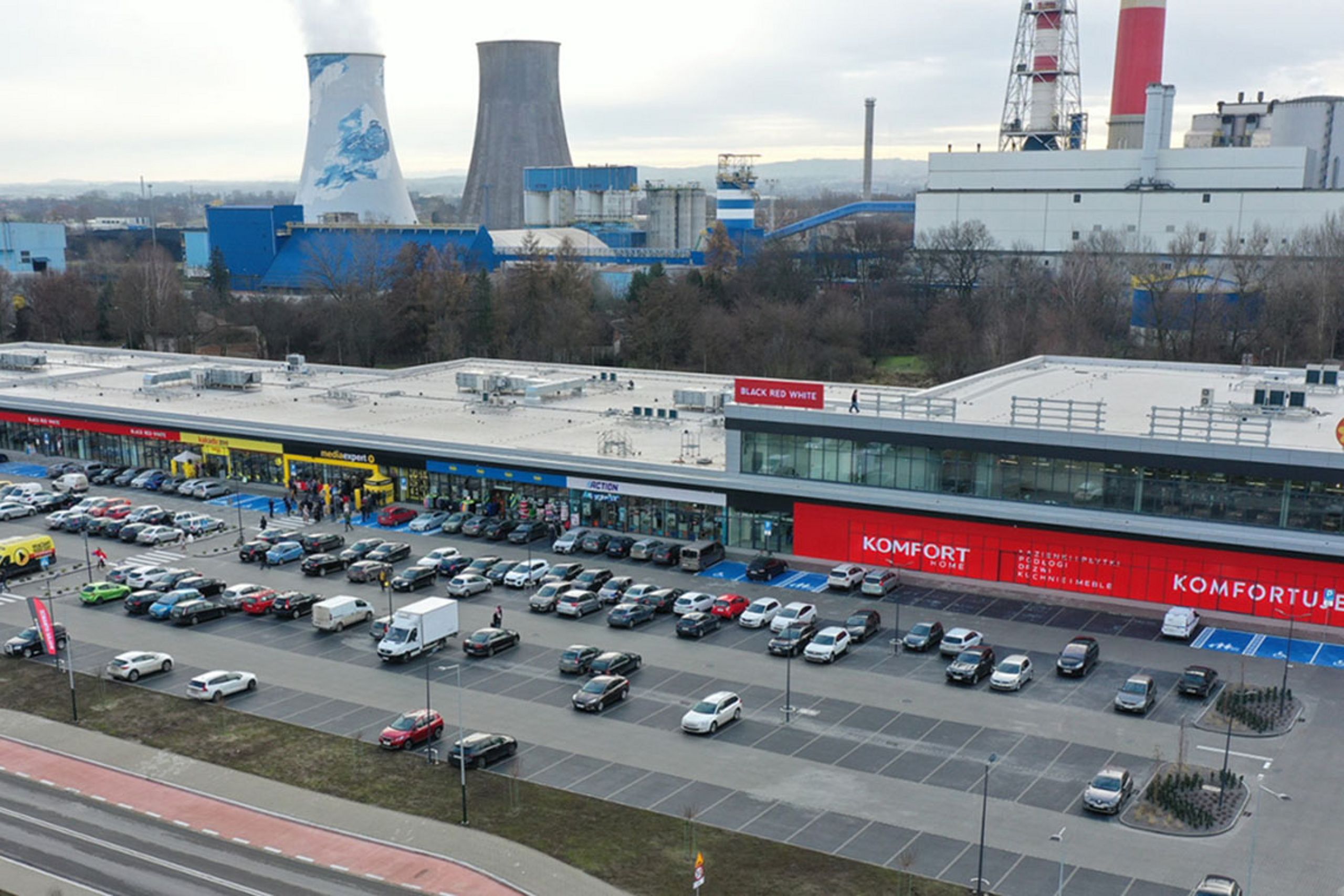 Widok z lotu ptaka na centrum handlowe z dużym parkingiem i elektrownią w tle.