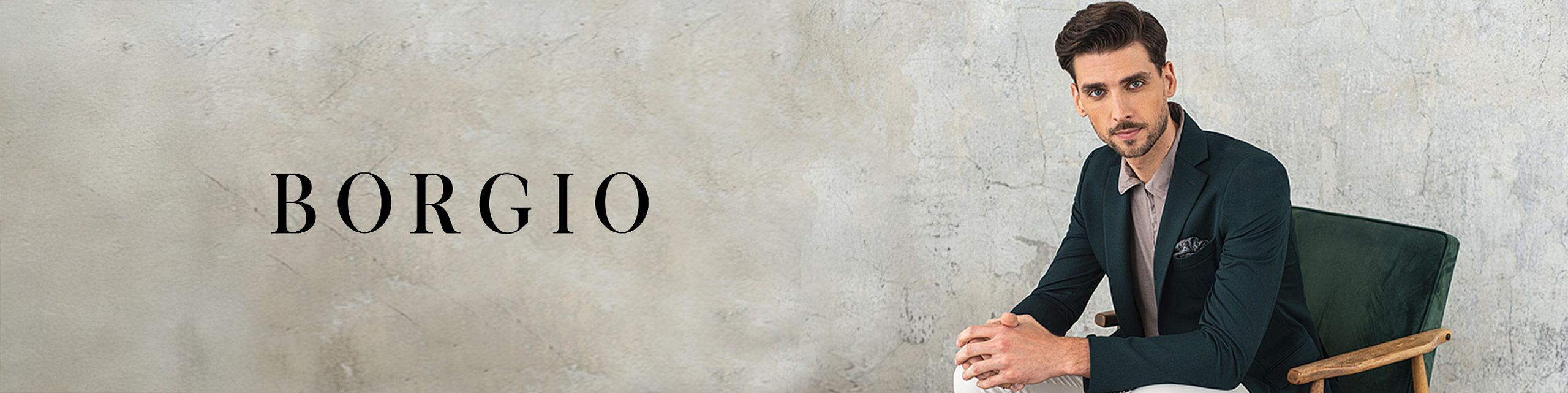 A stylish man wearing a dark green blazer and a light shirt sits on a chair against a light concrete wall. On the left side, the black text “BORGIO” – the fashion brand's logo – is displayed.