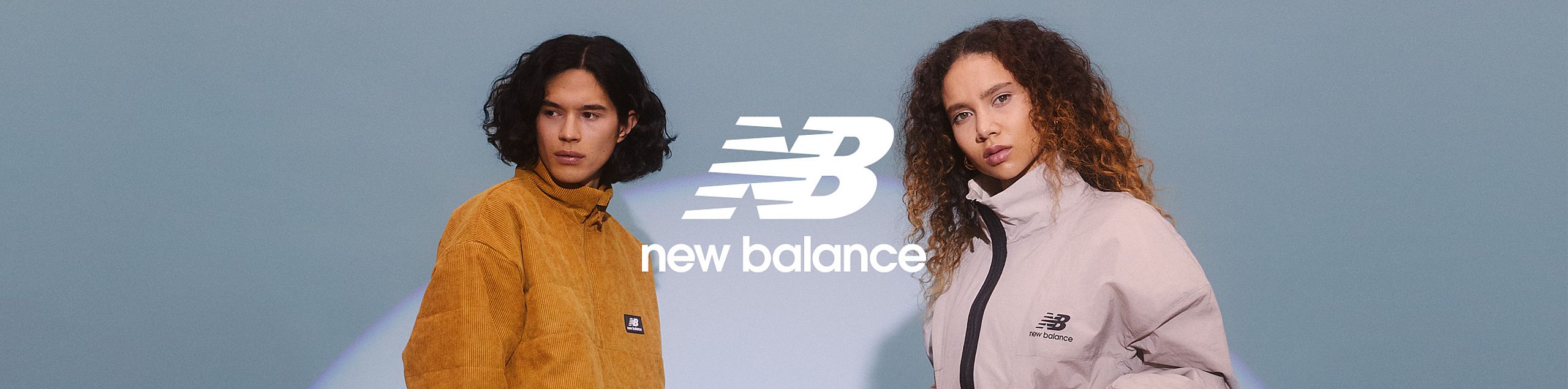 Dwoje młodych modeli w sportowych kurtkach marki New Balance – mężczyzna w musztardowej, kobieta w jasnoszarej – pozuje na tle niebieskiego gradientu. W centrum widoczne jest duże, białe logo „NB” i napis „new balance”.