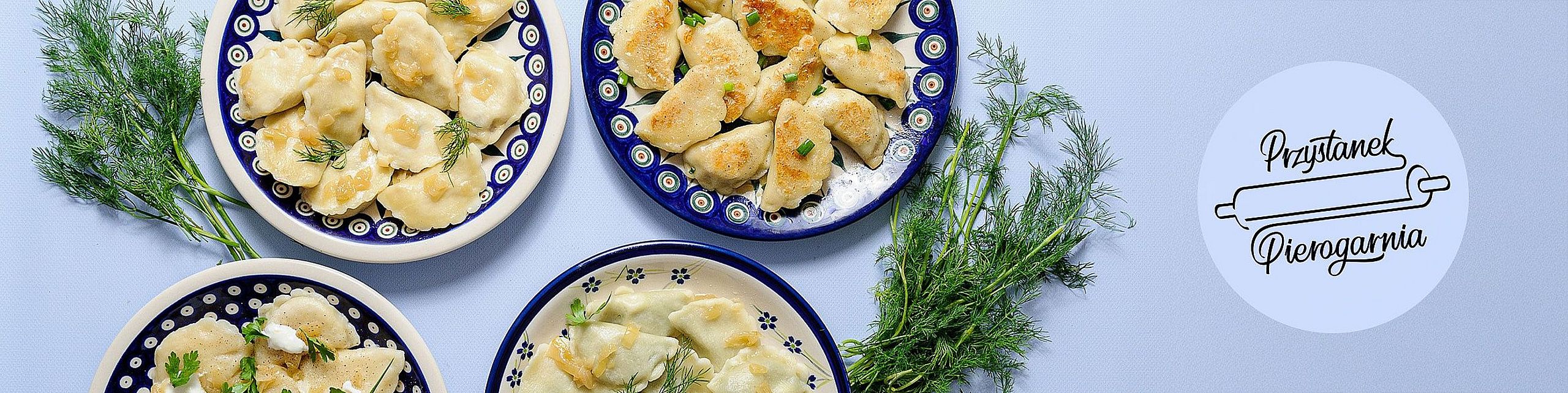 Cztery talerze wypełnione tradycyjnymi pierogami – gotowanymi i smażonymi – udekorowane świeżym koperkiem. Talerze ustawione na jasnym, niebieskim tle, obok gałązki koperku. Po prawej stronie okrągłe logo „Przystanek Pierogarnia” z grafiką wałka do ciasta.