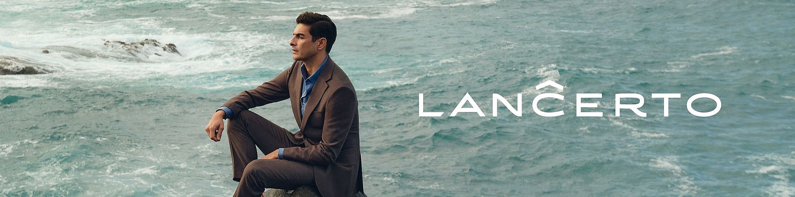 Mężczyzna w eleganckim, brązowym garniturze i niebieskiej koszuli siedzi na tle wzburzonego morza, wpatrzony w dal. Jego spokojna postawa kontrastuje z ruchem wody. Po prawej stronie widnieje białe logo „LANCERTO”.