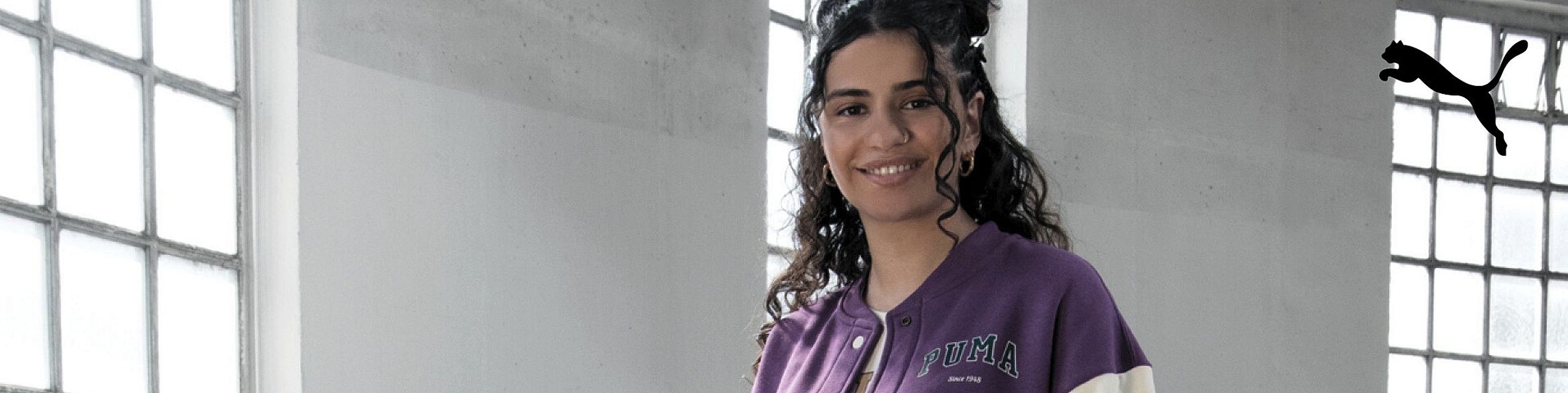 Uśmiechnięta kobieta z kręconymi włosami pozuje w jasnym pomieszczeniu przy dużych oknach, ubrana w fioletową bluzę Puma z logo na piersi. W prawym górnym rogu widoczne jest czarne logo Puma z sylwetką skaczącego kota.