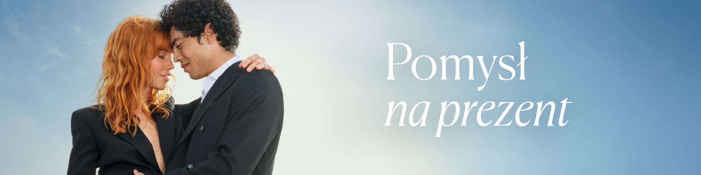 Para ubrana w eleganckie garnitury, stojąca blisko siebie i patrząca sobie w oczy w romantycznym uścisku. Na tle jasnego nieba widoczny jest napis "Pomysł na prezent". 