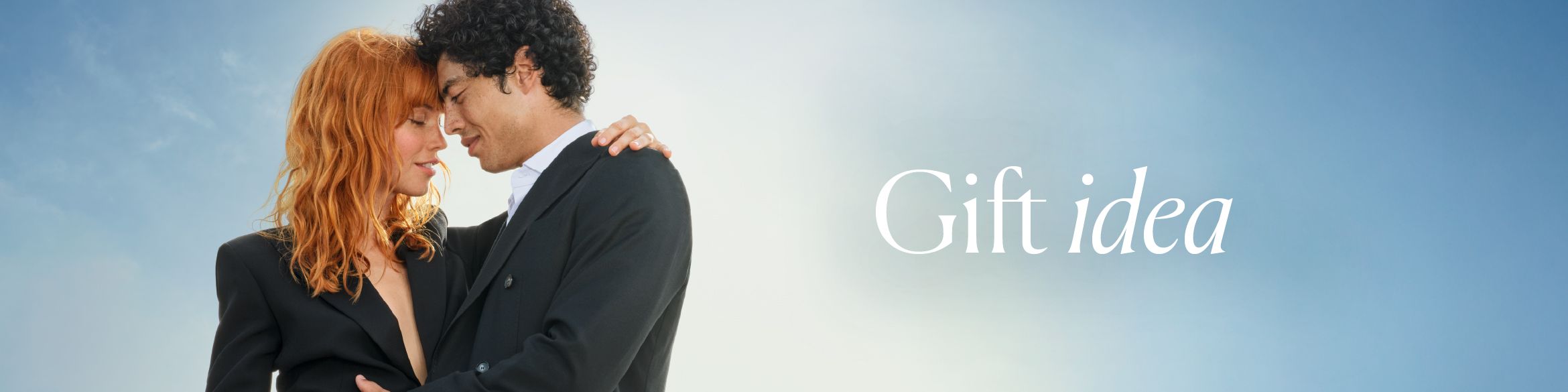 A couple dressed in elegant suits, standing close together and gazing into each other's eyes in a romantic embrace. In the background, the bright sky shows the text "Gift Idea".