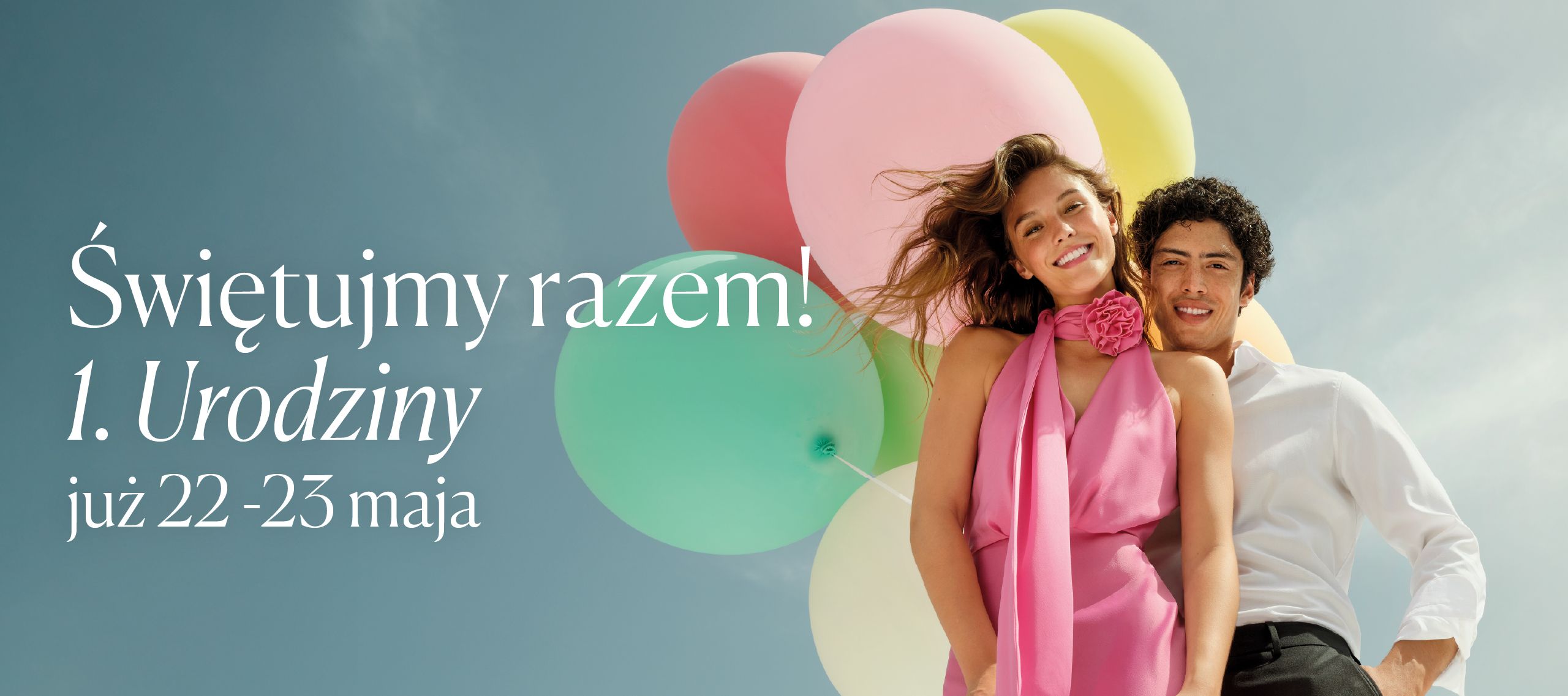 Para młodych ludzi na tle kolorowych balonów, uśmiechają się, celebrując pierwsze urodziny, z napisem 'Świętujmy razem! 1. Urodziny już od 22 maja'.
