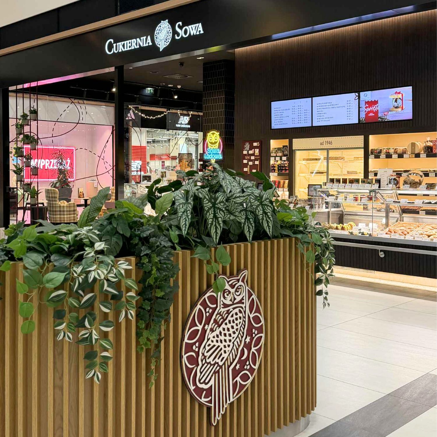 Entrance to Cukiernia Sowa in a shopping mall with an owl logo and green plants.