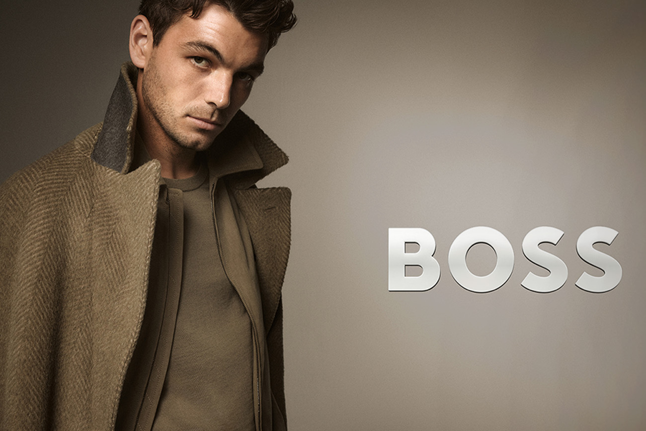 Mężczyzna w długim, eleganckim płaszczu w odcieniach brązu patrzy w stronę aparatu; po prawej stronie znajduje się logo ‘BOSS’.