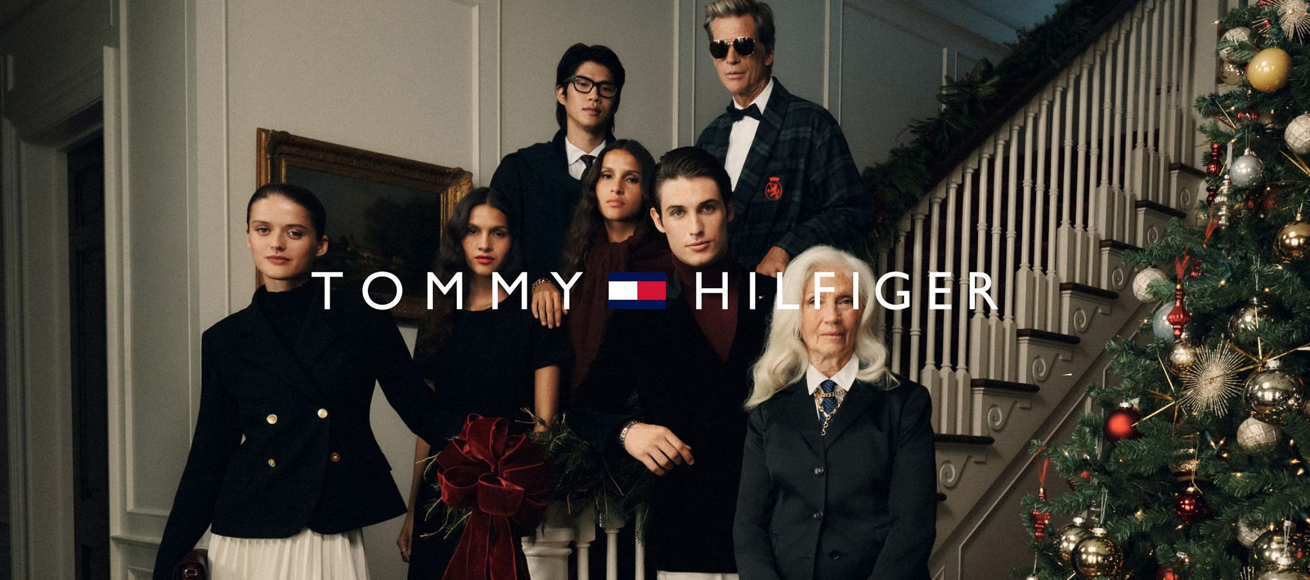 A group of elegantly dressed people pose inside a house next to a Christmas tree; Tommy Hilfiger holiday campaign.
