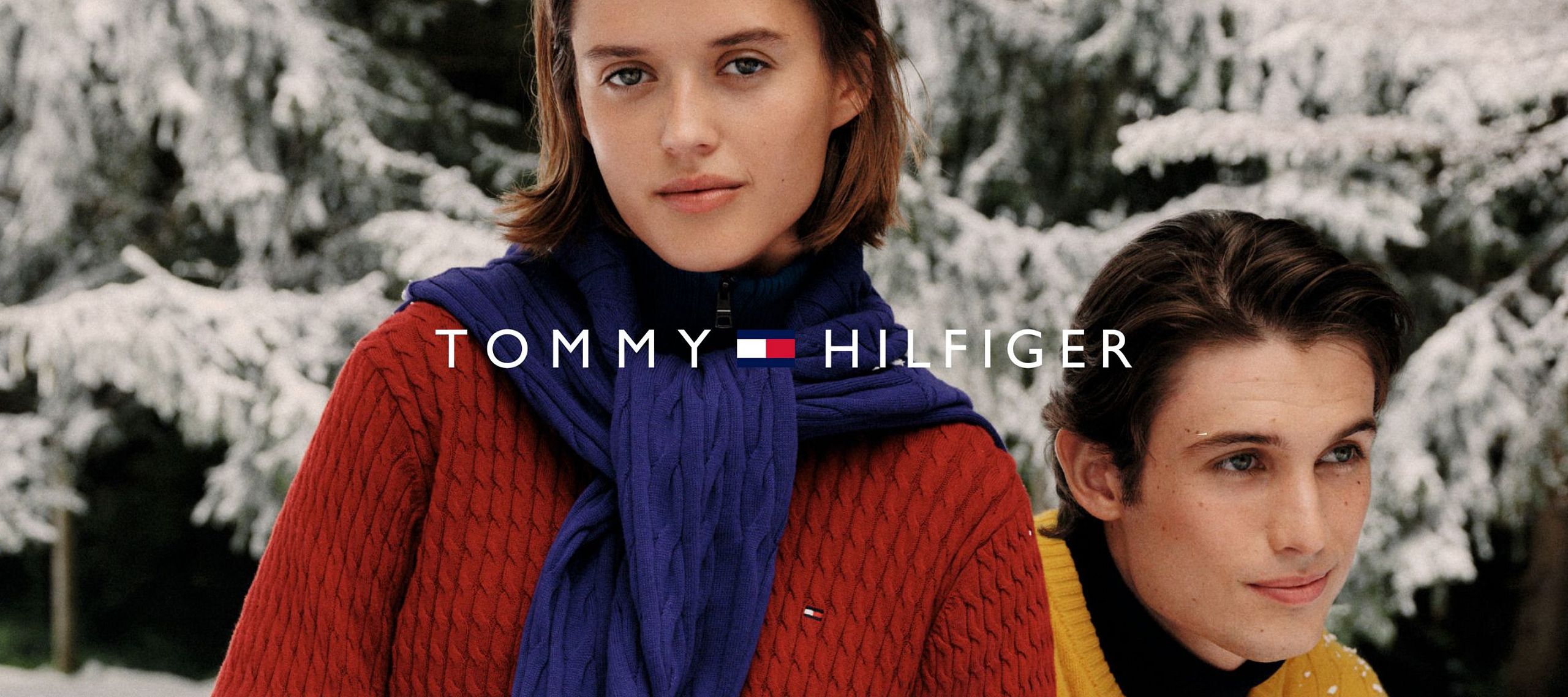 Dwie osoby w zimowych swetrach na tle zaśnieżonych drzew, logo Tommy Hilfiger.