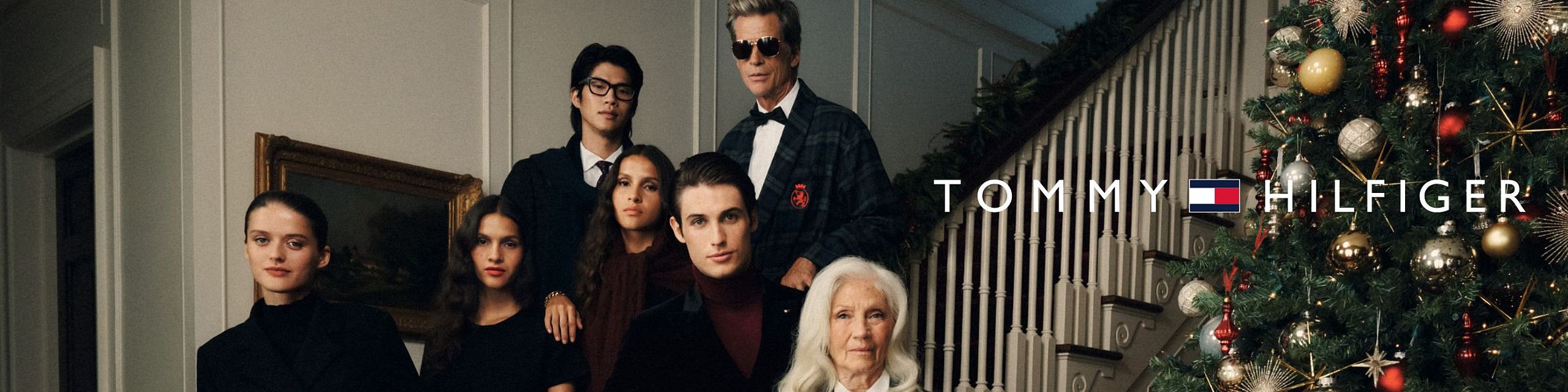 A group of elegantly dressed people pose inside a house next to a Christmas tree; Tommy Hilfiger holiday campaign.