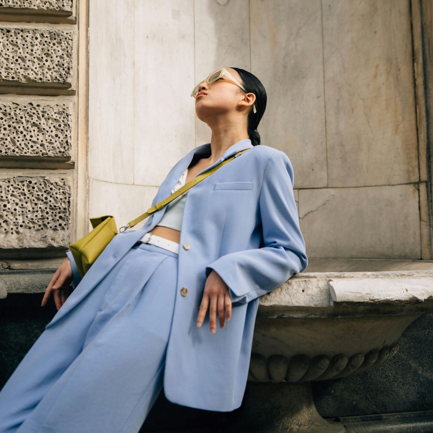Modelka w jasnoniebieskim garniturze i okularach przeciwsłonecznych oparta o kamienną fontannę.