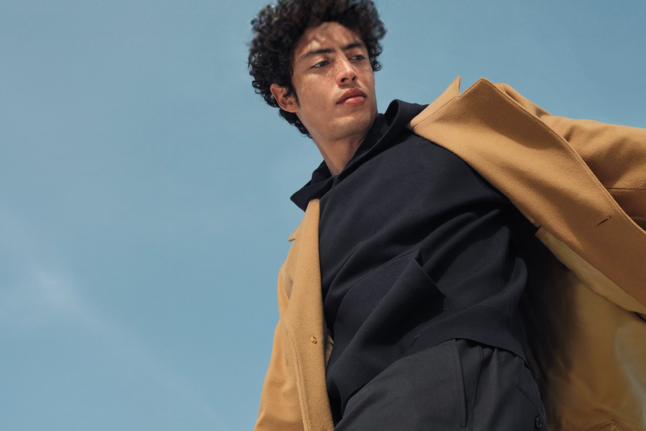 Mężczyzna w beżowym płaszczu i ciemnym swetrze pozuje na tle nieba – kampania modowa BOSS.