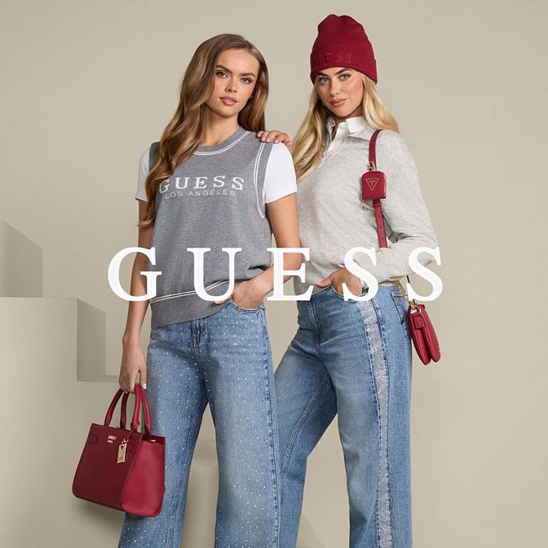 Dwie kobiety w casualowych stylizacjach z jeansami i szarymi bluzami Guess pozują na beżowym tle. Jedna trzyma czerwoną torebkę, druga ma na sobie czerwony beret i małą torebkę na ramię.