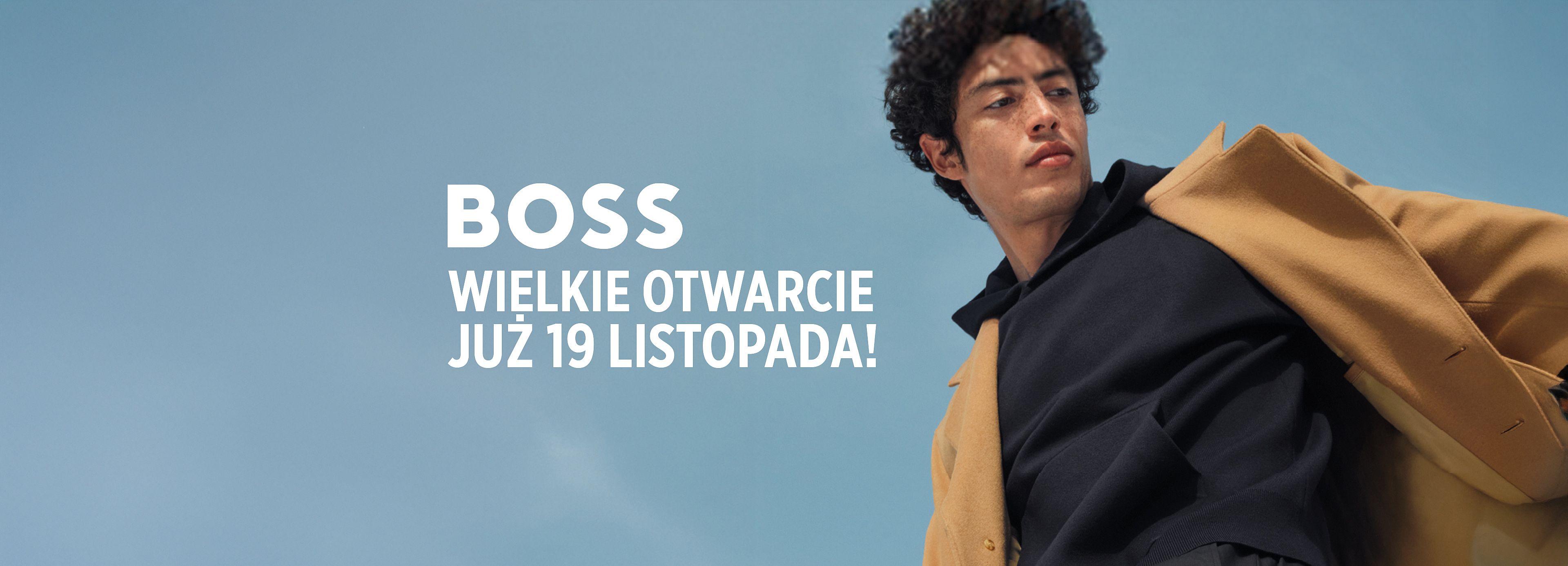 Mężczyzna w beżowym płaszczu i czarnej bluzie patrzy w bok na tle błękitnego nieba. Po lewej stronie widnieje napis: BOSS – Wielkie otwarcie już 19 listopada.