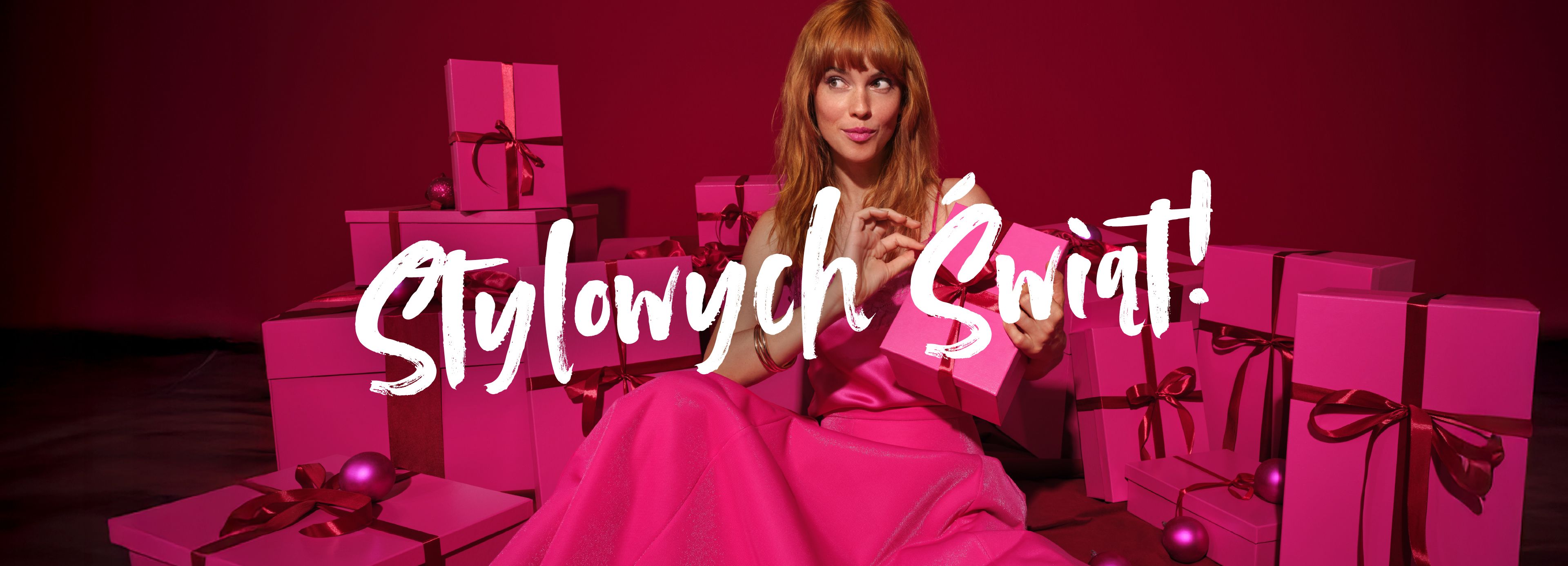 Kobieta w różowej sukience wśród różowych prezentów na czerwonym tle, z napisem ‘Stylowych Świąt!’