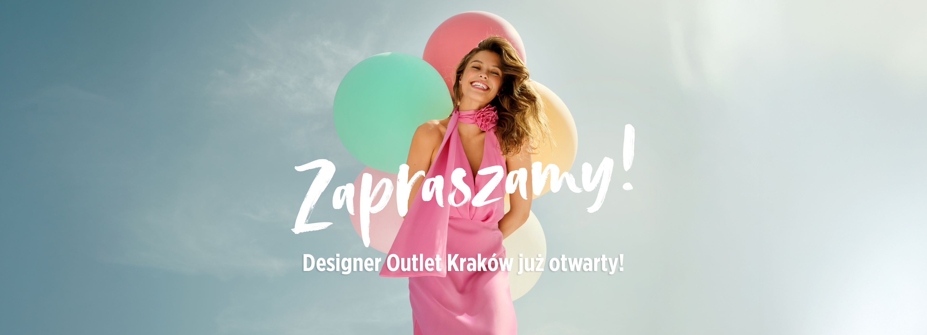 Uśmiechnięta kobieta w różowej sukience trzymająca pastelowe balony na tle błękitnego nieba. Napis: „Zapraszamy! Designer Outlet Kraków już otwarty!”
