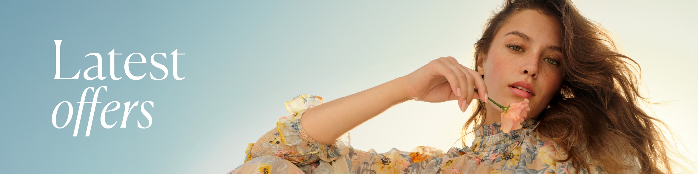 A woman in a light, floral dress holding a delicate pale pink flower near her face.