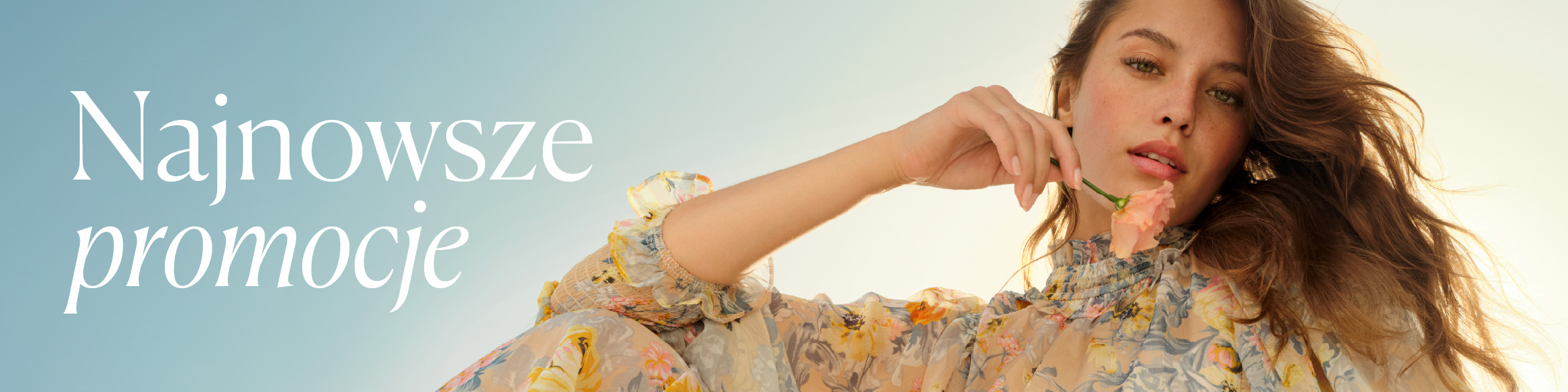 Kobieta w zwiewnej, kwiecistej sukience trzyma przy twarzy delikatny, jasnoróżowy kwiat.