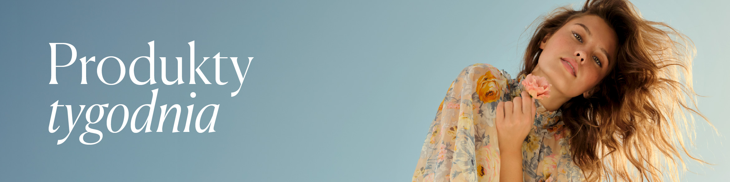 Kobieta w zwiewnej, kwiecistej sukience trzyma przy twarzy delikatny, jasnoróżowy kwiat.