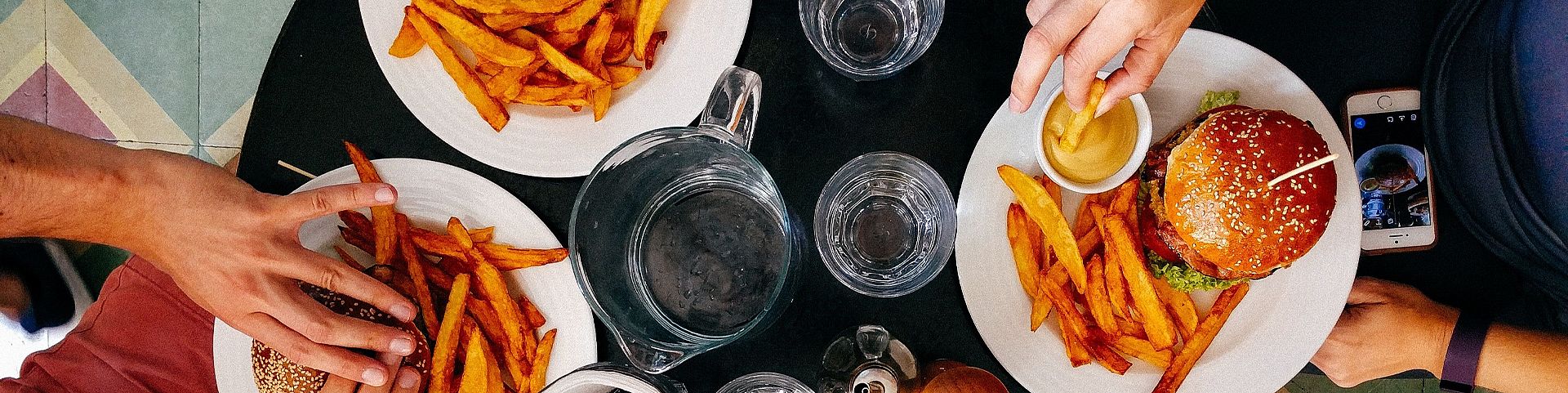 Three people sitting at a table eating a meal – burgers with fries and a dish of grilled vegetables. On the table are glasses of water, a pitcher, ketchup, beer, and condiments. Top-down view.