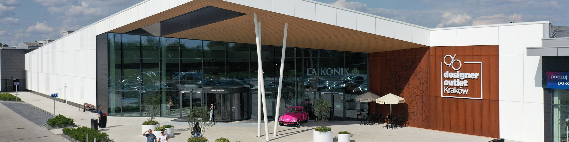 Frontowe wejście do Designer Outlet Kraków z nowoczesną architekturą i dużymi przeszklonymi ścianami. Nad drzwiami obrotowymi widoczny jest napis "Witamy / Welcome". Po lewej stronie znak TAXI, po prawej logo outletu oraz kawiarnia La Konik z różowym samochodem retro i parasolami. Przed wejściem ludzie siedzą i spacerują.